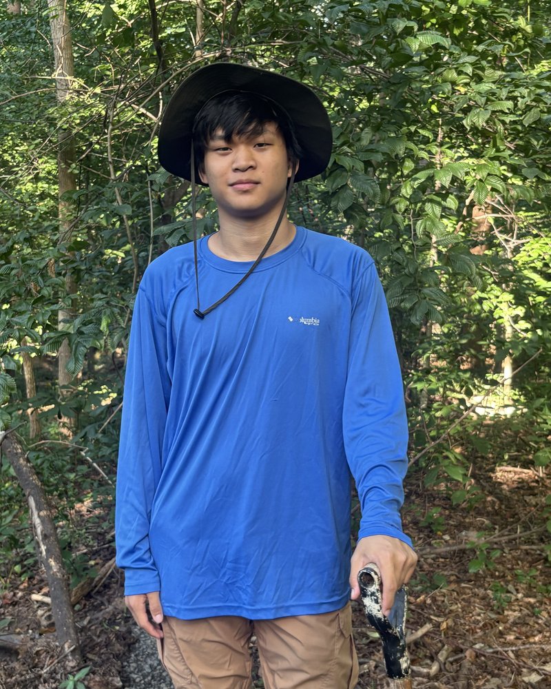 Daniel Tang on a woodland trail in a wide-brimmed hat and a
                blue long-sleeved shirt, framed from the hat to the waist.