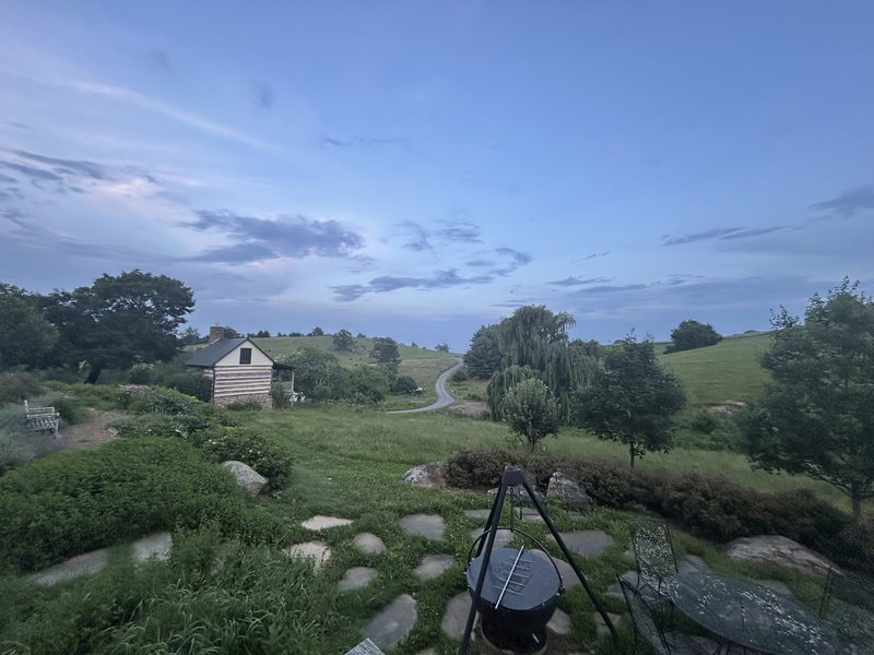 The log cabin and the winding farm road at dusk, wooded hills and a willow under a deep blue evening sky.