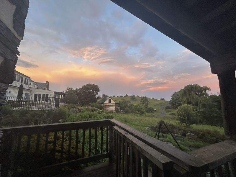 Looking out from a covered porch at sunset over rolling pasture: a farmhouse to the left, a small log cabin below, a willow, and a track running over the hill under pink and orange cloud.