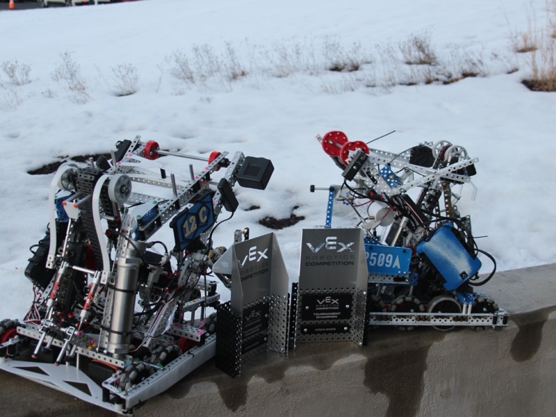 Two aluminum VEX robots posed on a wall in front of snow, two Tournament Champion trophies between them.