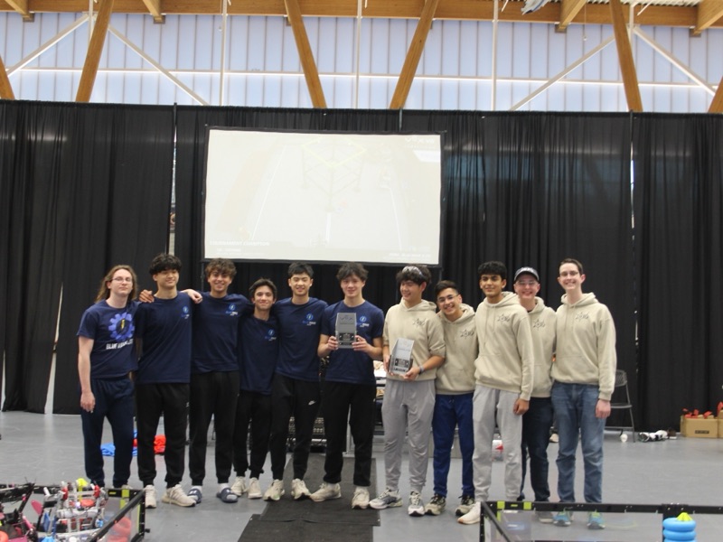 Eleven students from two teams lined up arm in arm between the fields, two holding VEX trophies, the screen behind reading Tournament Champion with Blue Wave Elite named.