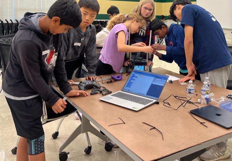 Six young students around a table mid-build, parts, zip ties and a laptop of block code spread between them.
