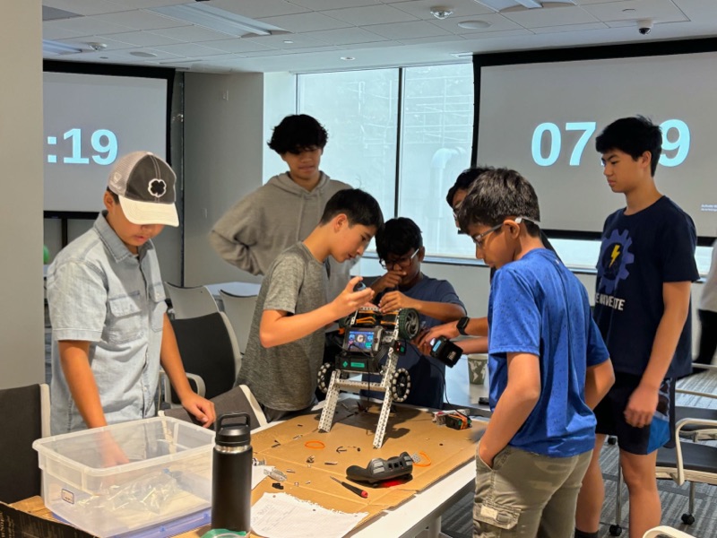Builders crowded around a half-assembled robot on a cardboard-covered table, one fitting a part while the others watch, countdown timers projected behind them.