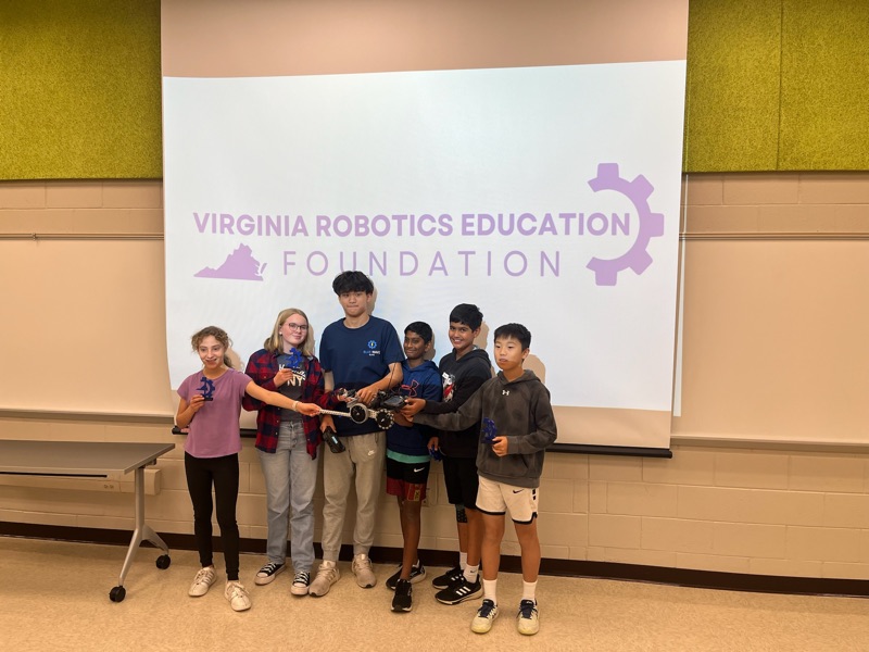 Workshop builders holding their finished robot and small trophies in front of a Virginia Robotics Education Foundation screen.