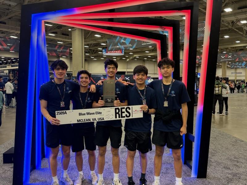 Five team members with medals holding the 2509A Blue Wave Elite team plate and a trophy under a lit arch at the VEX World Championship.
