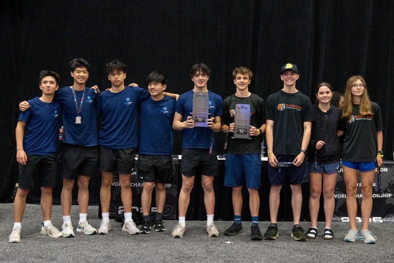 Nine students from the two alliance teams lined up arm in arm in front of the black World Championship backdrop, two silver VEX trophies held up between them.