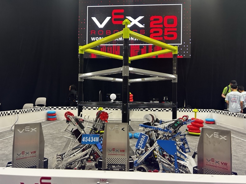 Two competition robots, 45434M and 2509A, parked on the field with three VEX trophies, under a screen reading VEX Robotics 2025 World Championship.